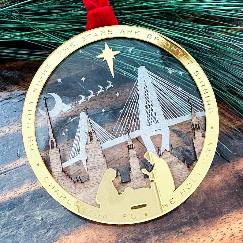 May include: A gold-colored Christmas ornament with a laser-cut design featuring a bridge, church steeples, and a nativity scene. The ornament has the words "OH HOLY NIGHT. THE STARS ARE BRIGHT! SHINING. CHARLESTON SC. THE HOLY CITY" around the edge.
