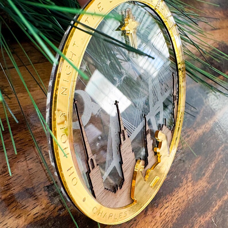 May include: A gold and clear Christmas ornament with the text "O Holy Night The Star of Bethlehem Charleston" encircling the edge. The ornament features a cityscape design with a star and bridge.