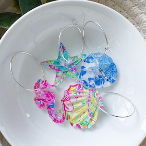 May include: A collection of colorful, patterned hoop earrings featuring sea-themed charms. The charms include a starfish, seahorse, seashell, and a round charm with a floral design. The earrings are set in a white bowl.
