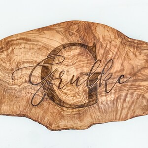 May include: A natural wood serving board with a unique, irregular shape. The board is engraved with the name "Grutke" and a large letter "G".