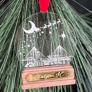 May include: A clear acrylic Christmas ornament featuring a white silhouette of the Charleston, SC skyline with a crescent moon and stars. The ornament is attached to a wooden base with a gold plaque that reads "Charleston, SC".