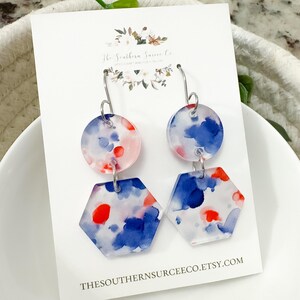 May include: Dangling earrings with a watercolor design. Each earring features a clear, round top and a clear, hexagonal bottom. The design includes blue, red, and pink watercolor splashes. The earrings hang from silver-colored hooks. The card behind the earrings says THESOUTHERNSURCEECO.ETSY.COM.
