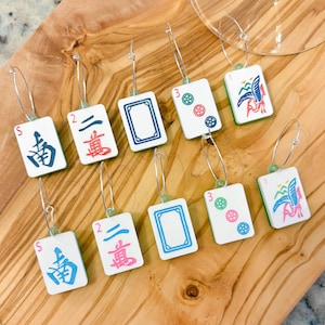 May include: A collection of Mahjong tile earrings with silver hoops. The earrings feature various tile designs with colorful symbols and Chinese characters. The earrings are displayed on a wooden surface.