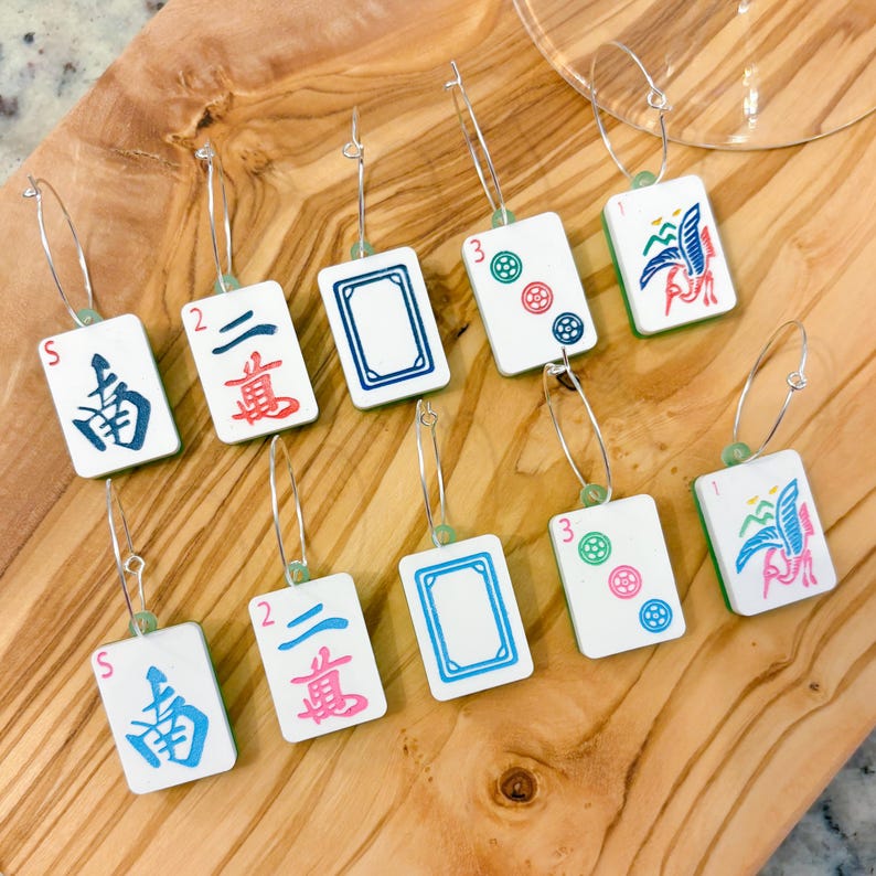 May include: A collection of Mahjong tile earrings with silver hoops. The earrings feature various tile designs with colorful symbols and Chinese characters. The earrings are displayed on a wooden surface.