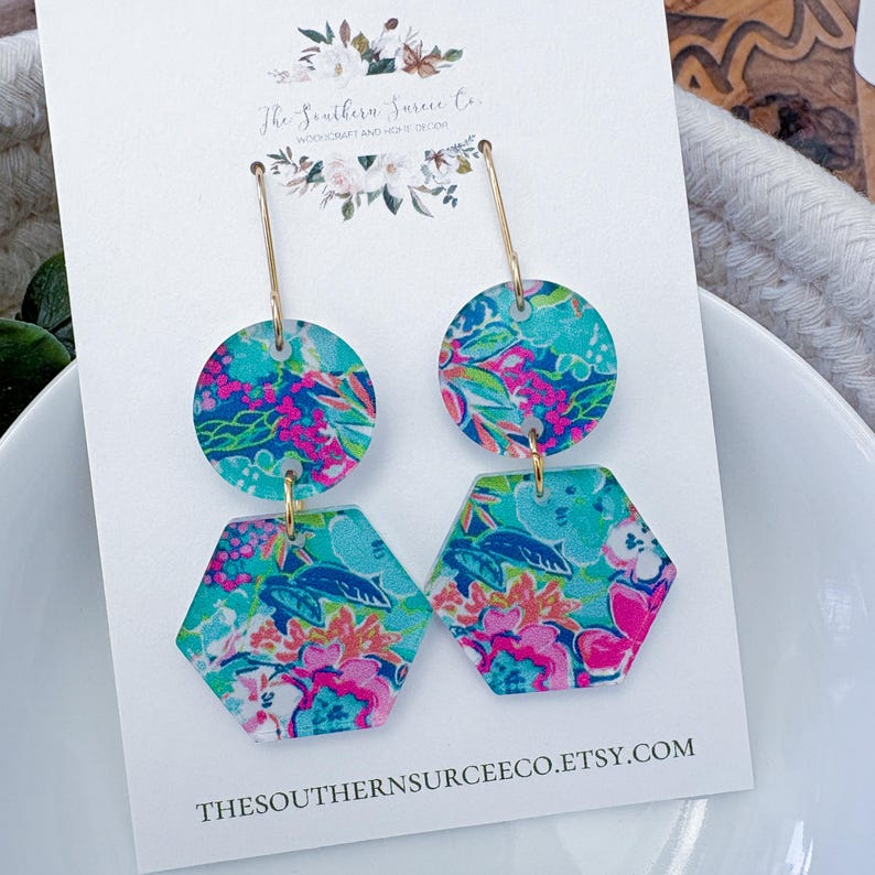 May include: A pair of dangle earrings with a floral pattern in turquoise, pink, and orange. The earrings have a gold-colored metal hook and are composed of a circle and a hexagon shape. The earrings are displayed on a white card with text.