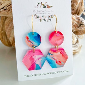 May include: A pair of dangle earrings with a colorful, abstract design. Each earring features a round top and a hexagonal bottom, connected by a gold-colored hook. The earrings have a vibrant mix of pink, blue, and orange hues. The text "The Southern Surcee Co." is visible.