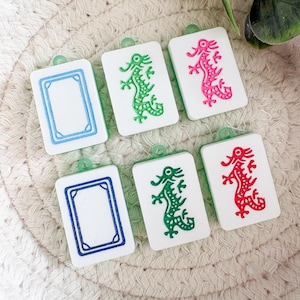 May include: Six white rectangular charms with green, pink, and red dragon designs and blue borders. Each charm has a small green loop at the top. The charms are arranged on a woven, beige surface.