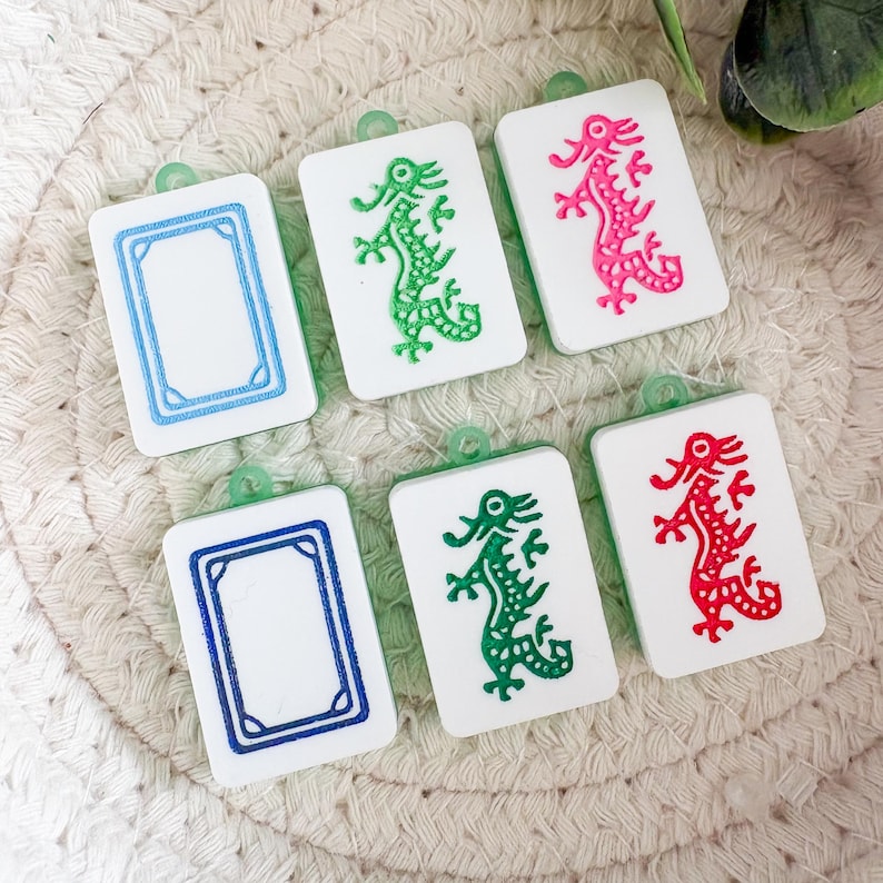 May include: Six white rectangular charms with green, pink, and red dragon designs and blue borders. Each charm has a small green loop at the top. The charms are arranged on a woven, beige surface.