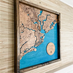 May include: A wooden map of Charleston, South Carolina, with a blue background and a wooden frame. The map is raised and has a 3D effect. The map is labeled "Charleston Harbor" in a circle.