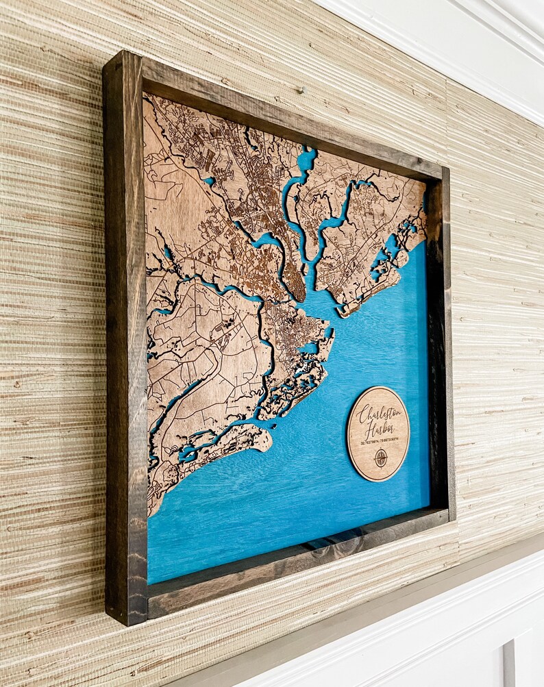 May include: A wooden map of Charleston, South Carolina, with a blue background and a wooden frame. The map is raised and has a 3D effect. The map is labeled "Charleston Harbor" in a circle.