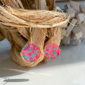 May include: A pair of gold hoop earrings with pink, green, and blue patterned round charms. The charms have a floral design.