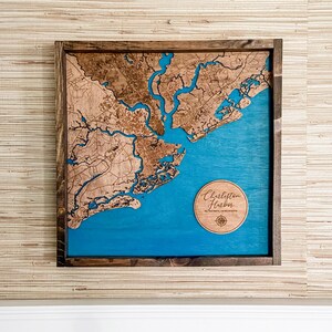 May include: A wooden map of Charleston Harbor, South Carolina, with a blue water feature and a compass rose. The map is framed in a dark wood frame and is hanging on a wall with a beige textured wallpaper.