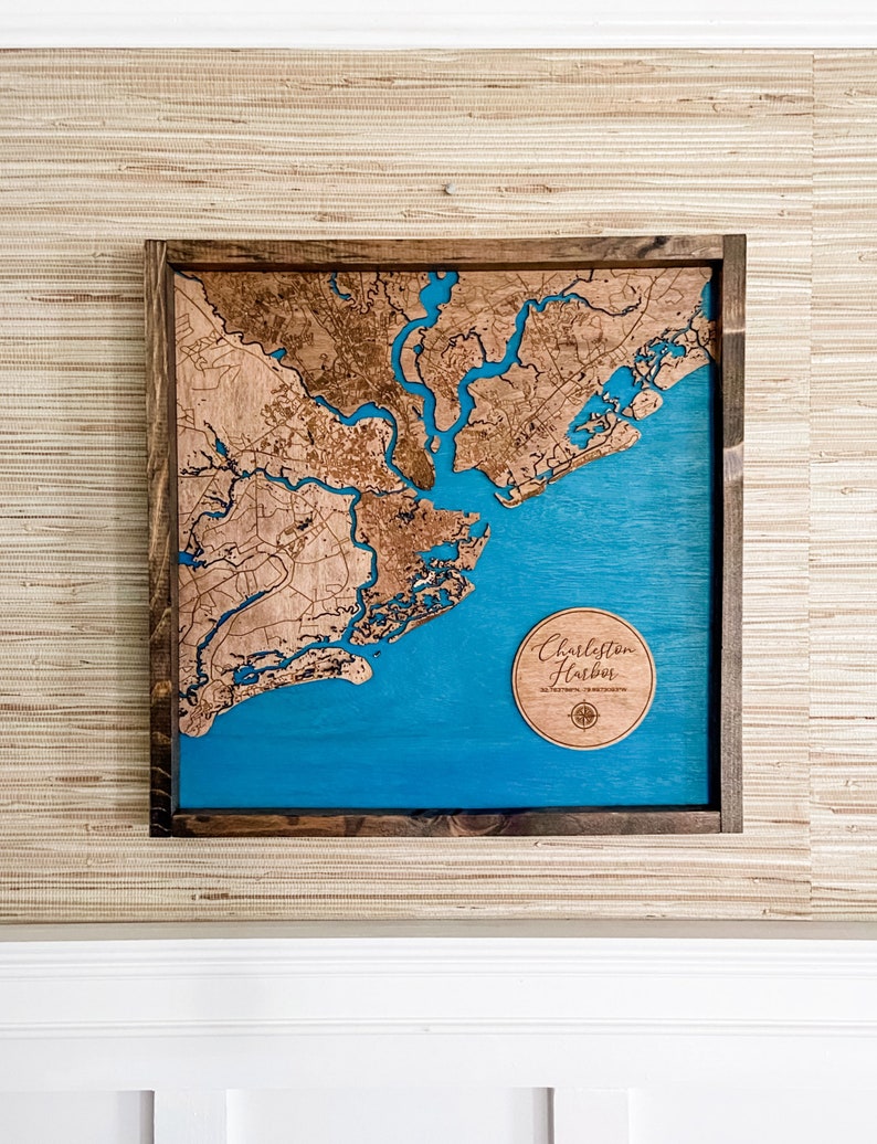 May include: A wooden map of Charleston Harbor, South Carolina, with a blue water feature and a compass rose. The map is framed in a dark wood frame and is hanging on a wall with a beige textured wallpaper.