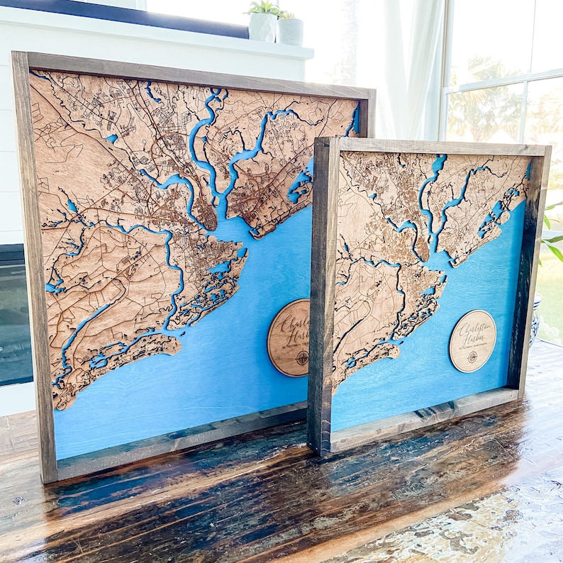 May include: Two wooden framed maps of Charleston Harbor, South Carolina. The maps are laser cut and have a blue background. The larger map is on the left and the smaller map is on the right. Both maps have a circular inscription that reads "Charleston Harbor".