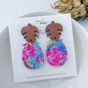 May include: A pair of earrings featuring a pink and blue floral design with a brown wooden leaf charm. The earrings are displayed on a white card with the text "thesouthernsourceco.etsy.com".