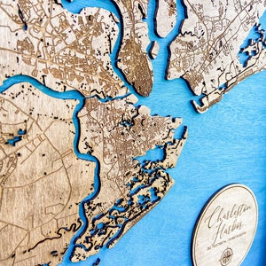 May include: A wooden map of Charleston Harbor, South Carolina, with a blue background. The map is laser-cut and shows the city's streets and waterways. A circular wooden plaque with the text "Charleston Harbor" and coordinates is in the lower right corner.