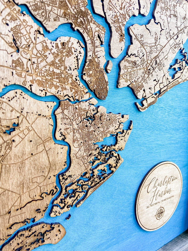 May include: A wooden map of Charleston Harbor, South Carolina, with a blue background. The map is laser-cut and shows the city's streets and waterways. A circular wooden plaque with the text "Charleston Harbor" and coordinates is in the lower right corner.