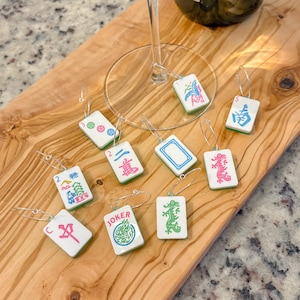 May include: A collection of white Mahjong tile wine charms with various symbols and numbers in blue, pink, and green. Each charm has a silver wire loop for attaching to a wine glass. The charms are displayed on a wooden cutting board.