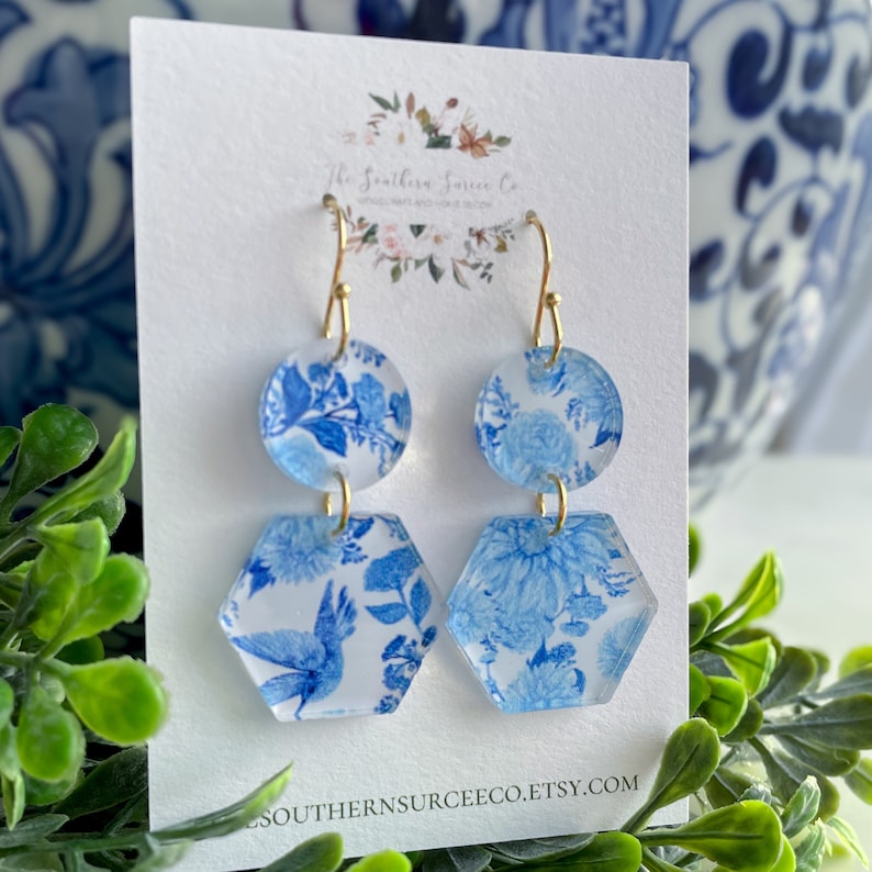 May include: Blue and white floral pattern earrings. The earrings have a gold hook and are made of two connected pieces: a circle and a hexagon. The earrings have a floral design and are on a white background.