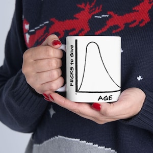 Puede incluir: Taza de cerámica blanca con un diseño gráfico negro. El gráfico muestra una curva etiquetada "F#CKS to Give" en el eje vertical y "AGE" en el eje horizontal. La taza es sostenida por una persona que lleva un suéter azul marino con un patrón de renos rojos.