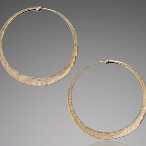 Pair of hammered solid 14k gold hoop earrings with endless closure displayed against a neutral gray background. Medium-sized hoops feature a textured, hand-hammered surface that creates subtle facets catching light for a warm golden shimmer. The clean circular design showcases the premium craftsmanship with slightly flattened profile and secure clasp. Warm gold tone provides luxurious alternative to silver version.