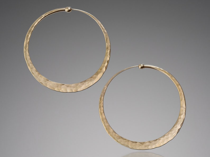 Pair of hammered solid 14k gold hoop earrings with endless closure displayed against a neutral gray background. Medium-sized hoops feature a textured, hand-hammered surface that creates subtle facets catching light for a warm golden shimmer. The clean circular design showcases the premium craftsmanship with slightly flattened profile and secure clasp. Warm gold tone provides luxurious alternative to silver version.