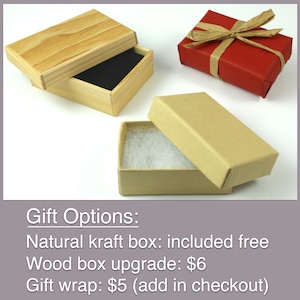 Gift packaging options photo showing three jewelry box styles. Natural kraft box (open to show black interior lining and cotton padding) and closed kraft box displayed alongside a red gift-wrapped box with bow. Purple text banner below explains options: "Gift Options: Natural kraft box: included free, Wood box upgrade: $6, Gift wrap: $5 (add in checkout)." Perfect for showing customers packaging presentation for gifting jewelry.