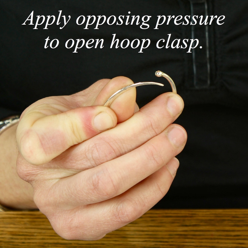Close-up instructional image showing hands demonstrating how to open gold hoop earring. Person's thumb and index finger apply opposing pressure to open the clasp, with the gold hoop held against a wood grain surface. Text overlay reads "Apply opposing pressure to open hoop clasp." Image clearly shows the technique for proper handling of the endless hoop design for easy wear and removal.