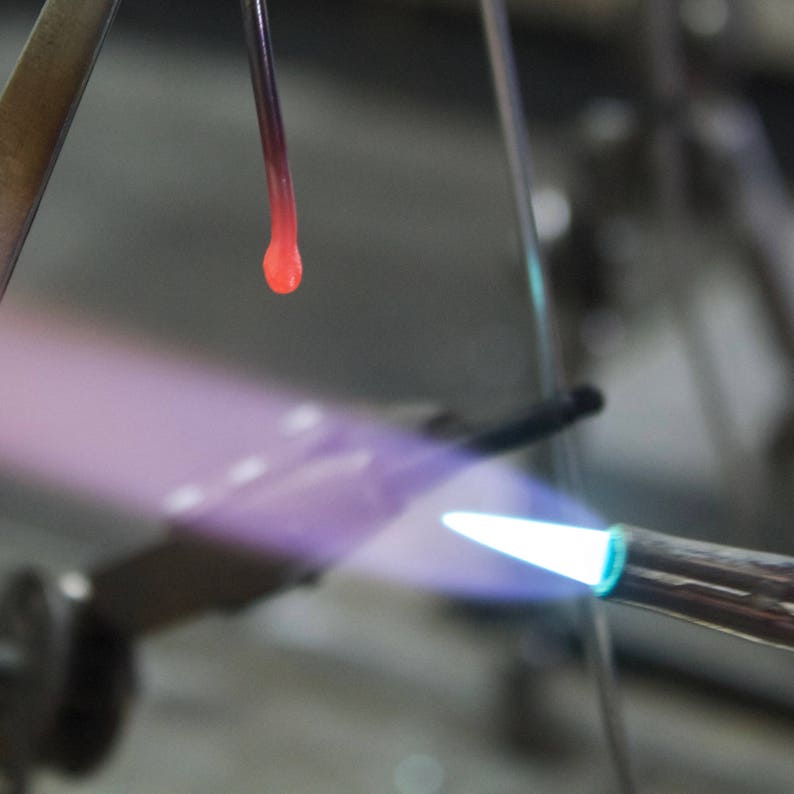 Close-up image of metalsmithing process showing intense flame melting metal into a glowing orange-red droplet. Torch flame appears blue at the tip where it contacts the metal. Part of a soldering setup visible with tools in soft focus background. Demonstrates the high-temperature techniques used to create handcrafted gold jewelry in the artist's studio. Shows the craftsmanship behind each handmade piece.