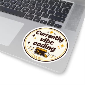 May include: A circular sticker with a cream background and a dark brown border. The text reads Currently vibe coding in dark brown letters. It features a small orange computer terminal icon with a cursor and several small yellow star accents.
