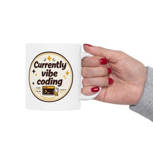 May include: A white ceramic mug featuring a circular graphic with the text Currently vibe coding in dark brown letters. The design includes a small orange computer window icon with a cursor and several small yellow star accents on a cream background.