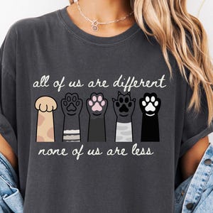 May include: Dark gray t-shirt with the text "all of us are different, none of us are less." The shirt features a graphic of five different colored paw prints. The shirt has short sleeves and a relaxed fit.