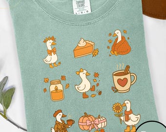 May include: A light green Comfort Colors t-shirt featuring a collection of fall-themed illustrations. The design includes cartoon ducks with autumn accessories, pumpkin pie, a candle, a mug of hot cocoa, pumpkins, and fall leaves. The word "Bay" is in a circle.