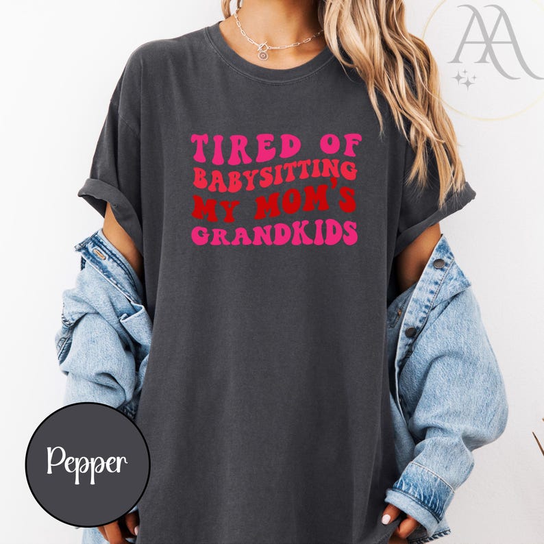 May include: Dark gray t-shirt with the text "TIRED OF BABYSITTING MY MOM'S GRANDKIDS" in pink and red retro font. The shirt is worn with a denim jacket. The overall theme is casual wear.