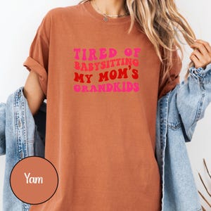 May include: A rust-colored t-shirt with the text "TIRED OF BABYSITTING MY MOM'S GRANDKIDS" in pink and red lettering. The shirt has rolled-up sleeves and a casual fit. A denim jacket is draped over the shoulders.