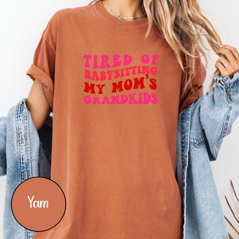 May include: A rust-colored t-shirt with the text "TIRED OF BABYSITTING MY MOM'S GRANDKIDS" in pink and red lettering. The shirt has rolled-up sleeves and a casual fit. A denim jacket is draped over the shoulders.