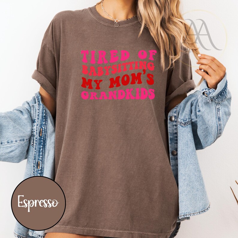 May include: Espresso-colored t-shirt with the text "TIRED OF BABYSITTING MY MOM'S GRANDKIDS" in pink and red. The shirt has rolled-up sleeves and is paired with a denim jacket.