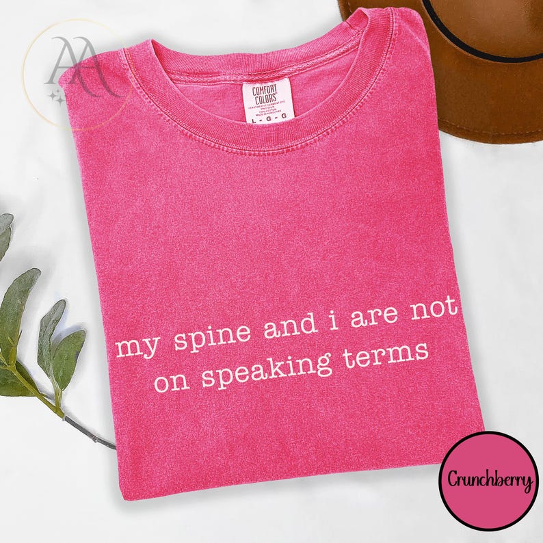 Pode incluir: Uma t-shirt rosa brilhante com o texto branco "my spine and i are not on speaking terms". A t-shirt tem gola redonda e uma etiqueta Comfort Colors. Um chap&eacute;u castanho &eacute; parcialmente vis&iacute;vel no fundo.