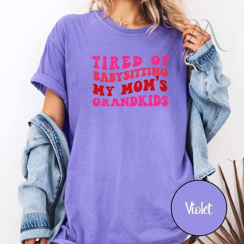 May include: Violet-colored t-shirt with the text "TIRED OF BABYSITTING MY MOM'S GRANDKIDS" in pink and red retro font. The shirt is a comfortable, casual style.