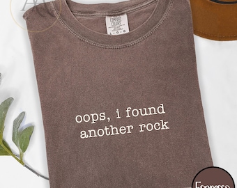 Puede incluir: Una camiseta de manga corta de color marrón oscuro con cuello redondo. La prenda presenta el texto en blanco "oops, i found another rock" impreso en el pecho. La etiqueta interior del cuello muestra la marca Comfort Colors y la talla L-G.