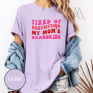 May include: A light purple t-shirt with the text "TIRED OF BABYSITTING MY MOM'S GRANDKIDS" in red and pink retro font. The shirt is a relaxed fit, with rolled-up sleeves. A denim jacket is draped over the shoulders.