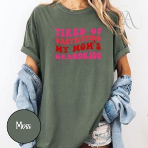 May include: Moss green t-shirt with the text "TIRED OF BABYSITTING MY MOM'S GRANDKIDS" in red and pink. The shirt has rolled-up sleeves and a relaxed fit. A denim jacket is draped over the shoulders.