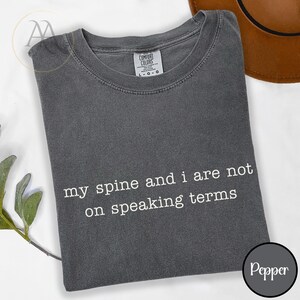 Pode incluir: T-shirt cinzenta escura com o texto branco "my spine and i are not on speaking terms". A camisa tem gola redonda e mangas curtas. A etiqueta da marca "Comfort Colors" &eacute; vis&iacute;vel.