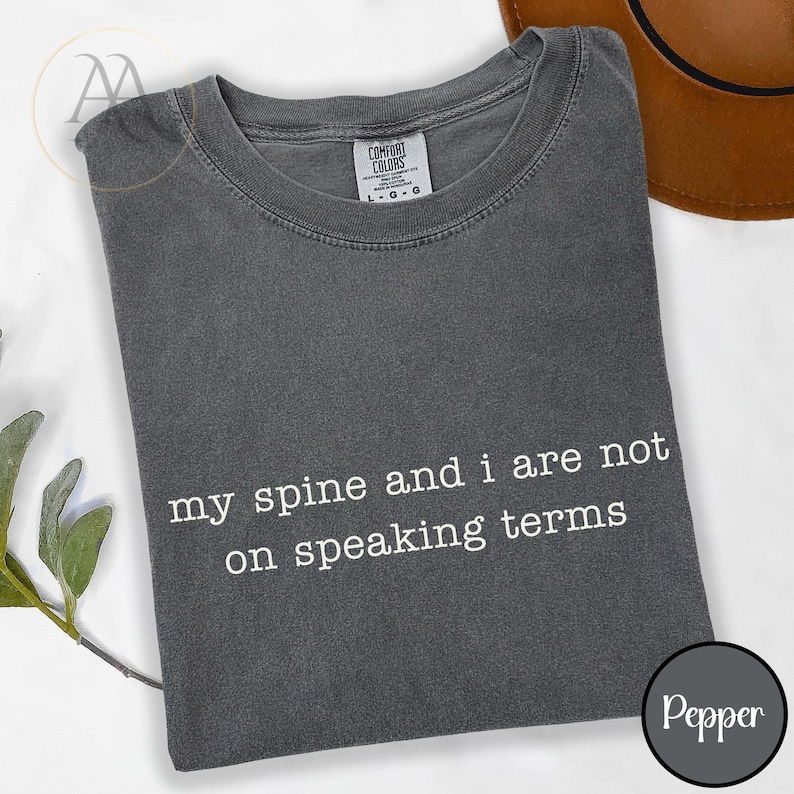 Pode incluir: T-shirt cinzenta escura com o texto branco "my spine and i are not on speaking terms". A camisa tem gola redonda e mangas curtas. A etiqueta da marca "Comfort Colors" &eacute; vis&iacute;vel.