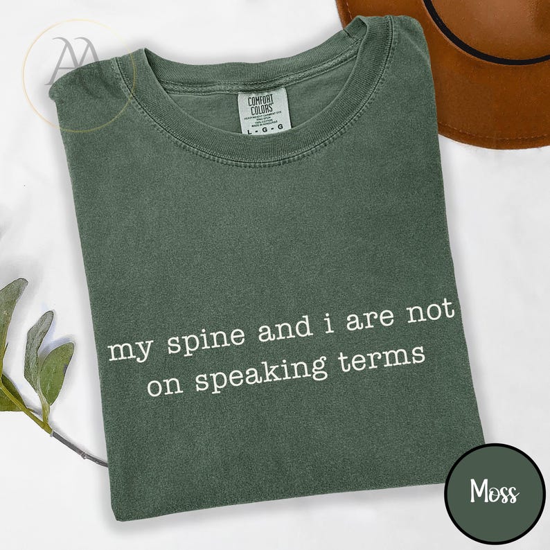 May include: A moss green t-shirt with the text "my spine and i are not on speaking terms" in white. The shirt has a crew neck and a tag that says "Comfort Colors". A brown hat is partially visible in the background.