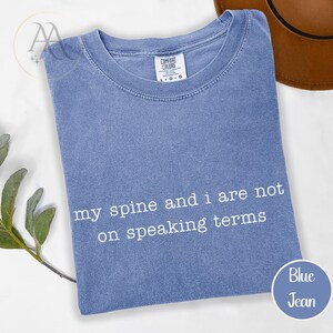 Pode incluir: Camiseta azul jeans com o texto branco "my spine and i are not on speaking terms". A camisa est&aacute; dobrada e tem uma etiqueta Comfort Colors. Um chap&eacute;u marrom &eacute; vis&iacute;vel no fundo.