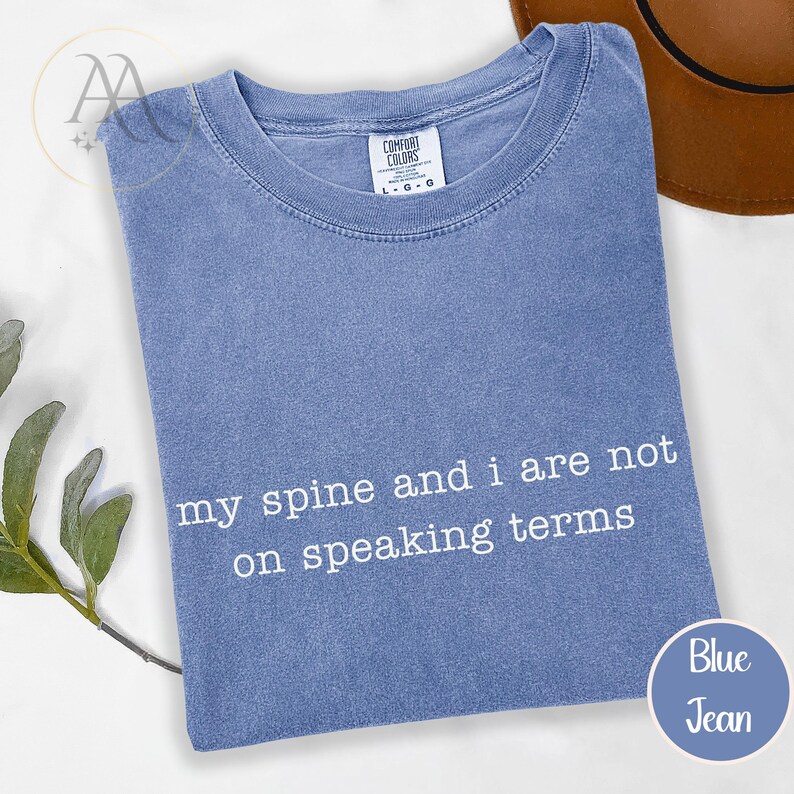 Pode incluir: Camiseta azul jeans com o texto branco "my spine and i are not on speaking terms". A camisa est&aacute; dobrada e tem uma etiqueta Comfort Colors. Um chap&eacute;u marrom &eacute; vis&iacute;vel no fundo.