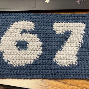 May include: A handmade, crocheted navy blue square with the numbers "67" in light gray. The numbers are prominently displayed in the center of the square, showcasing a contrasting color scheme and textured design.