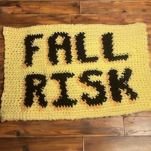 Puede incluir: Una alfombra rectangular de ganchillo hecha a mano en color amarillo claro. Las palabras "FALL RISK" están tejidas en negro, con un contorno color melocotón. La alfombra está colocada sobre un suelo de madera.
