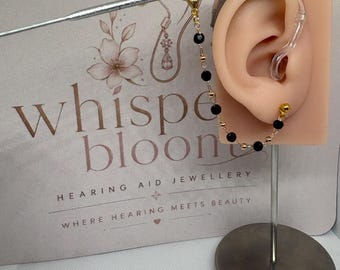 May include: A hearing aid charm featuring a delicate gold-tone chain interspersed with small black beads and gold-tone spacers. The chain attaches to a hearing aid worn on a beige ear model, which is displayed in front of a branded card with text.